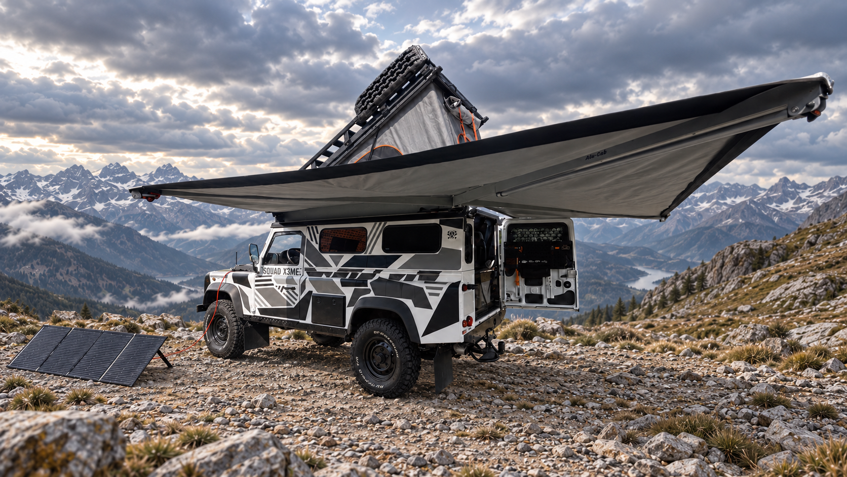 Der Defender im Camp-Aufbau in digital ergänzter felsiger Berglandschaft, mit geöffnetem Aufstelldach, weit ausladender Markise, offener Hecktür und mobilem Solarmodul.