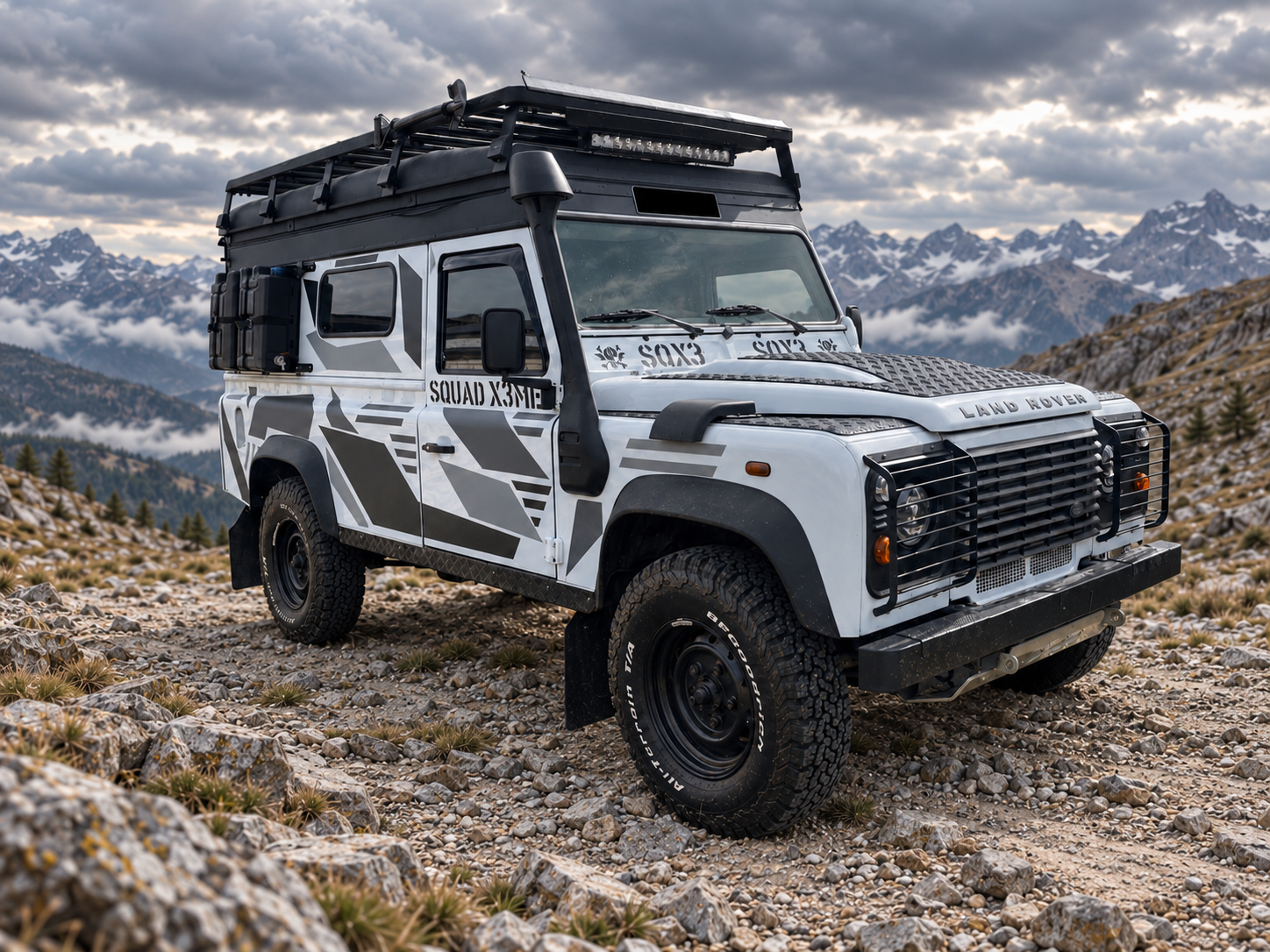 Der umgebaute Land Rover Defender von schräg vorn in digital ergänzter Felslandschaft: grauweißes X3ME-Design, schwarze Anbauteile, grobe Reifen und schmale LED-Leiste am Dach.