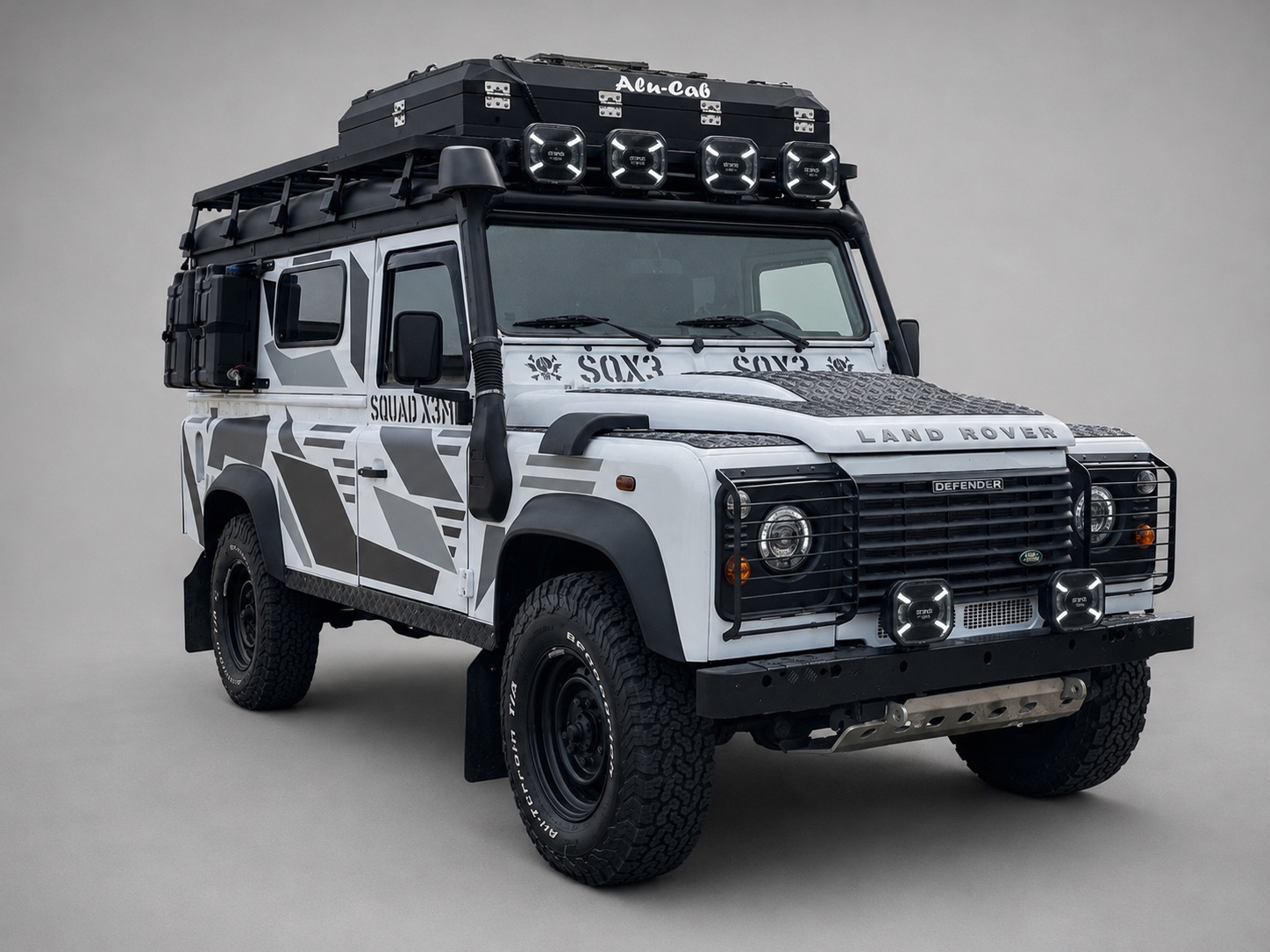Der umgebaute Land Rover Defender von schräg vorn: grauweißes X3ME-Design, schwarze Anbauteile, grobe Reifen und Zusatzscheinwerfer.